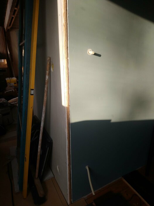 Renoveringsprojekt: dörr står, arbetsmaterial lutat mot vägg, strålande solljus, skuggor, oavslutad miljö.