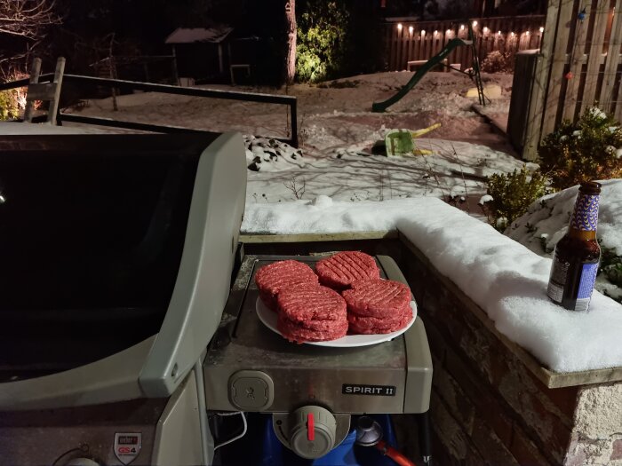 Grill med råa hamburgare, snöigt landskap bakom, ölflaska på kant, nattbelysning, vintergrillning utomhus.