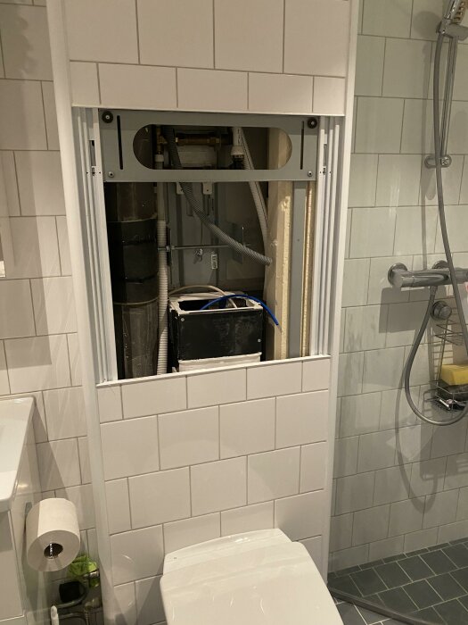 Ett öppet installationsutrymme ovanför en toalett i ett kaklat badrum med dusch. Rör och teknik synliga.