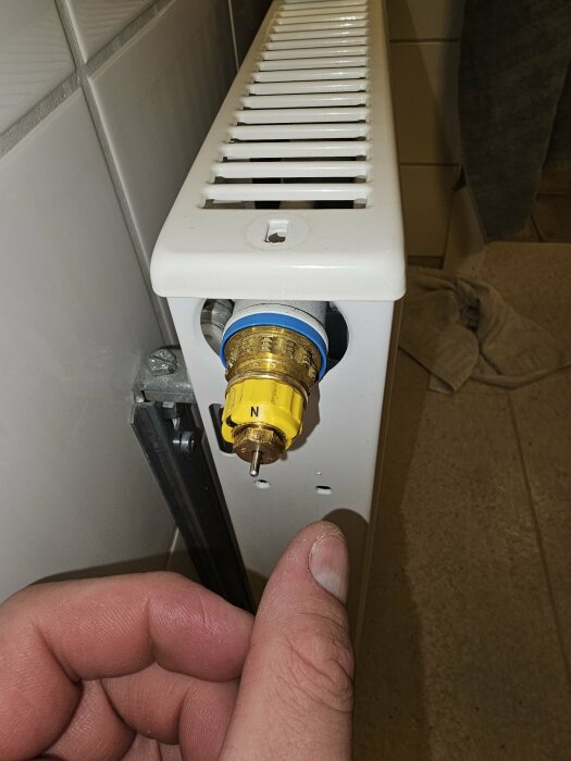 Hand justerar termostatventil på vit elementradiator. Gula och blåa delar synliga. Hemmiljö.