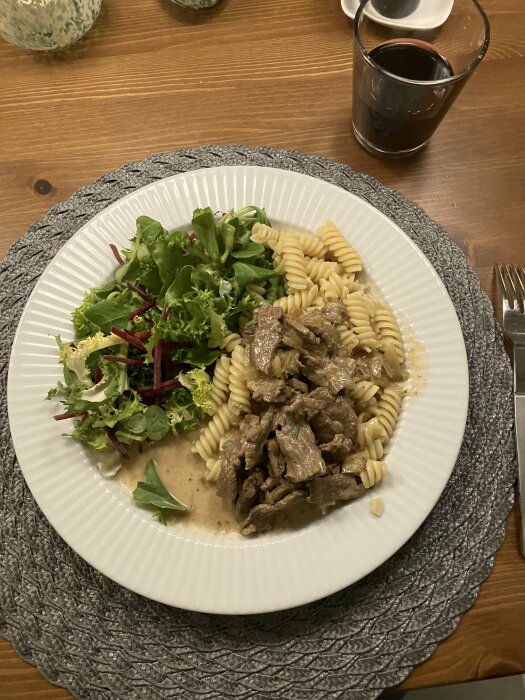 Ett middagsbord med pasta, gräddig sås, strimlat kött, sallad och ett glas mörk dryck.