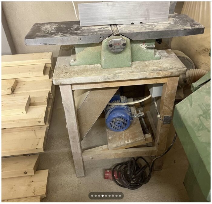 Verkstadsbild med sladdar, träbearbetningsmaskin och högar med träbitar. Dammit och använd utrustning syns.