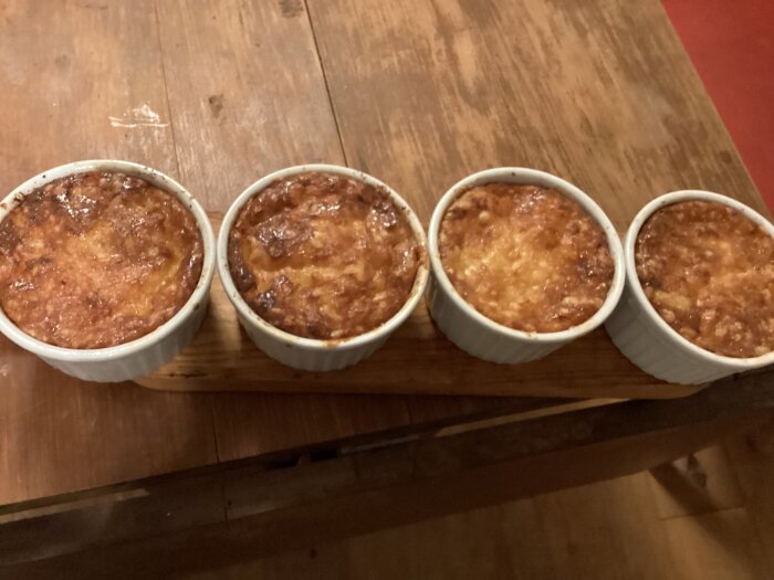Fyra gräddade maträtter i vita keramikskålar på träbord. Ser hemlagade och gyllenbruna ut.