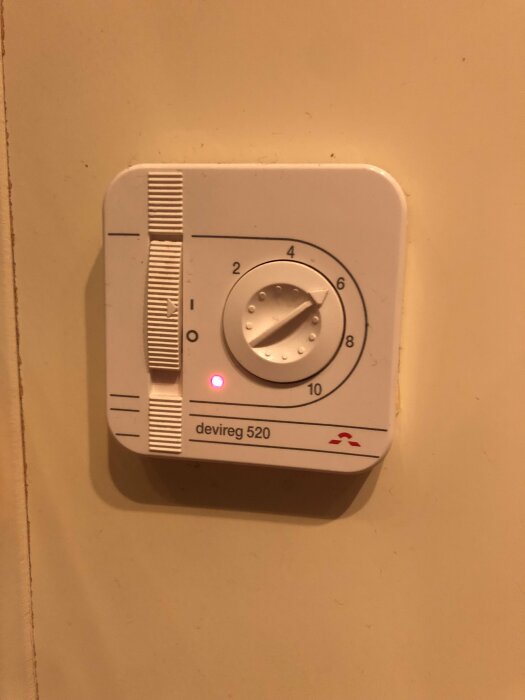 En termostat för uppvärmning, devireg 520-modellen, monterad på en vägg, med en tänd röd indikeringslampa.