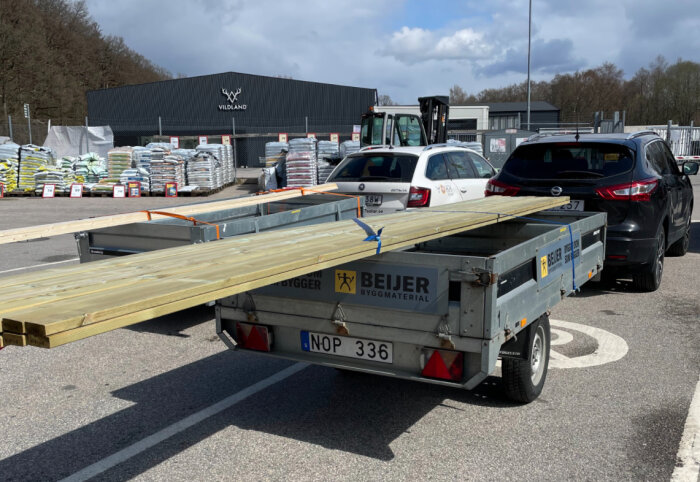 Parkeringsplats med släpvagn lastad med trä, fordon, bakom byggmaterialbutik. Säckar och gaffeltruck syns.