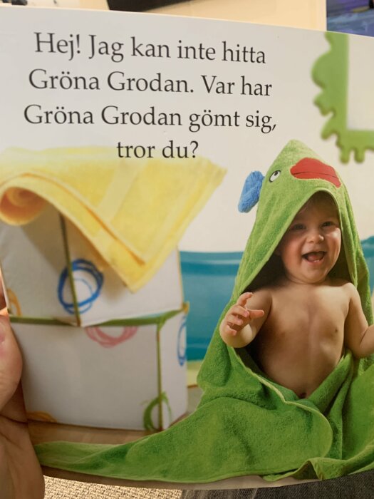 Ett barn i en grön grodhandduk ler, boktext frågar var "Gröna Grodan" är gömd.