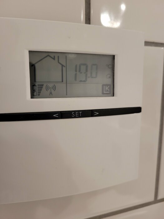 Digital termostat visar 19.0 grader Celsius, väggmonterad, inställningsknappar, husikon, trådlös signalikon, vit bakgrund.
