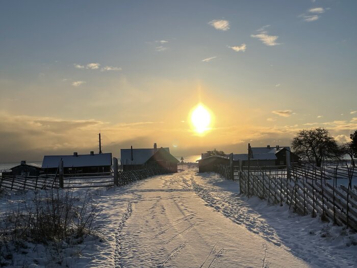 Solnedgång över snötäckt by med trästaket och hus, vinterlandskap, klar himmel, fotspår i snön.