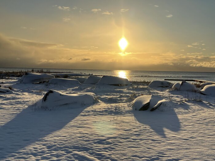 Solnedgång över snötäckt strand med uppochnervända båtar och reflektion över havet. Fredlig, vinter, skymning, kallt, stämningsfullt.