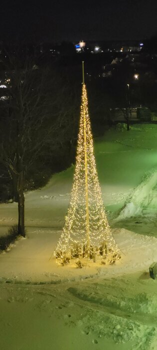 En stor upplyst julgran står i ett snötäckt landskap under natttid, med stadsbelysning i bakgrunden.