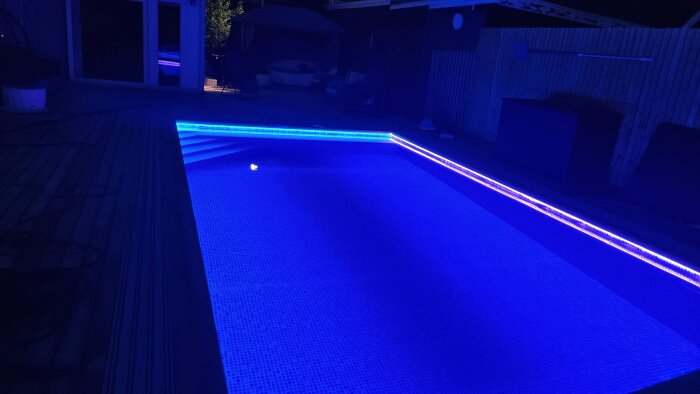 Utomhuspool med blått LED-ljus under kvällen, omgiven av terrass och trädgårdsmöbler.