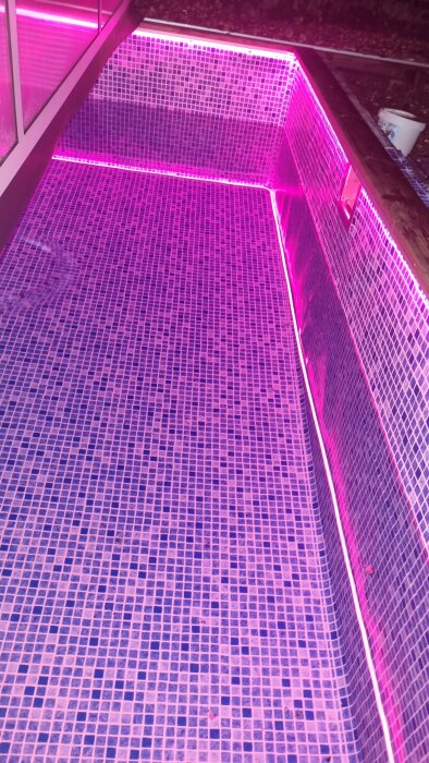 Ledbelysning belyser en tom swimmingpool med mosaikplattor, skapar ett neonrosa sken.