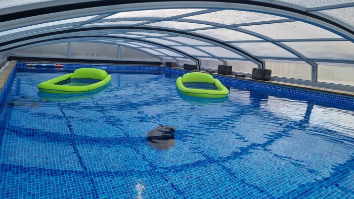 Simbassäng under genomskinligt tak med två gröna uppblåsbara ringar i vattnet. Lugnt och inbjudande.