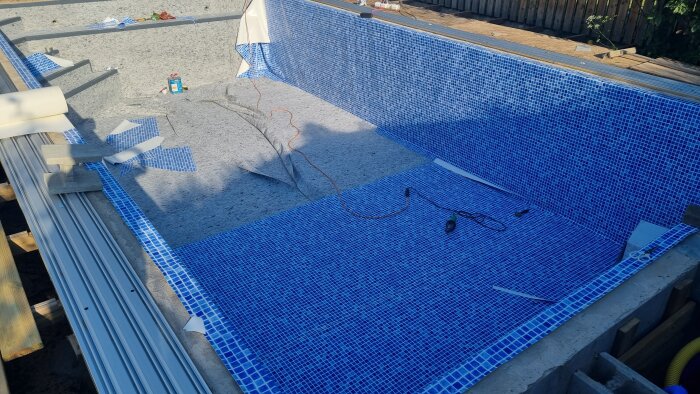 Pool under konstruktion med blå mosaikplattor, byggmaterial, och poolskydd vid sidan.