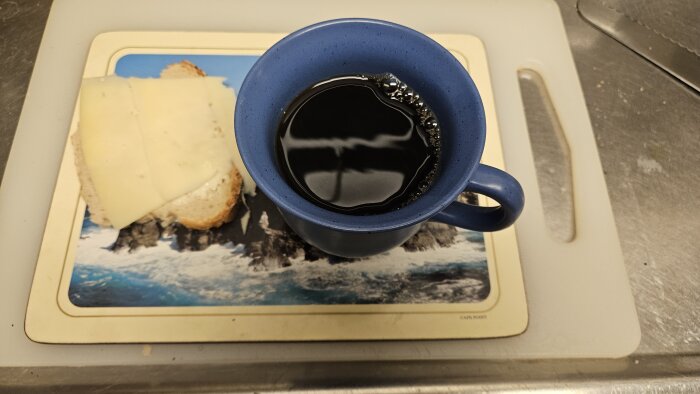 En kopp kaffe med en smörgås med ost på ett bergslandskapsbordunderlägg på en köksskärbräda.