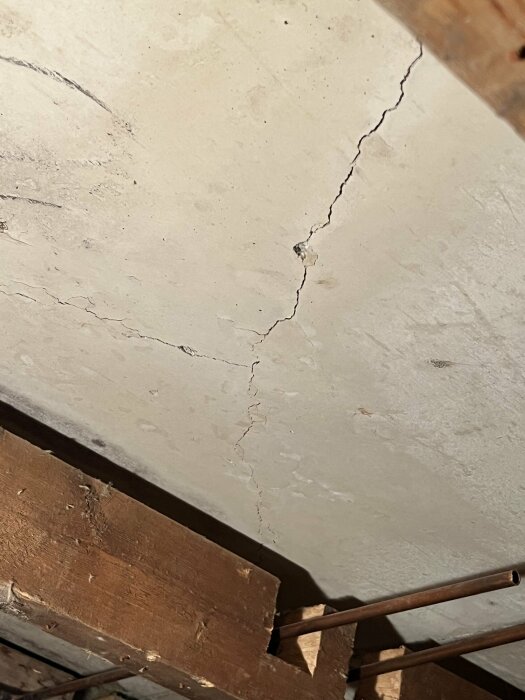 Sprucken vitmålad betongvägg över träbjälkar och metallstång. Byggnadsstrukturella defekter. Indirekt belysning. Skadad yta.