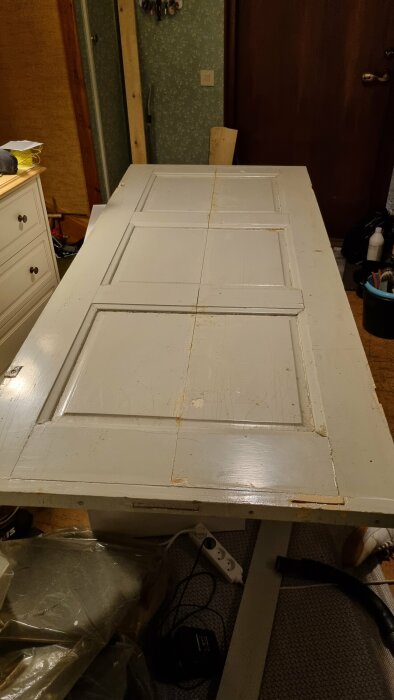 Vit dörr ligger horisontellt, används som arbetsyta, slarvigt målad, i rörigt inomhusutrymme.