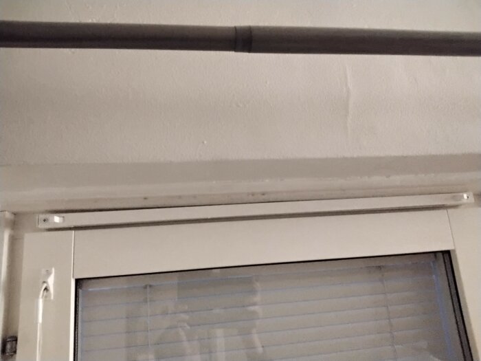 Ett fönster med persienner och en svart rörledning ovanför, inomhus, natt eller svagt upplyst rum.