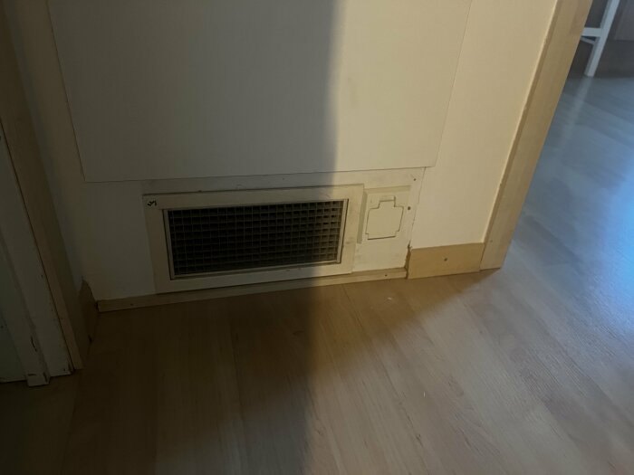 Vägg med ventilationsgaller och skadade delar nära golvet, inomhusmiljö med trägolv och dörröppning.