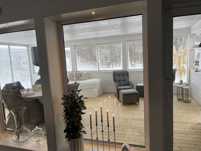 Inomhusvy med vitt vardagsrum, fönster mot snötäckt landskap, levande ljus förgrunden. Harmonisk, ljus, avslappnad atmosfär.