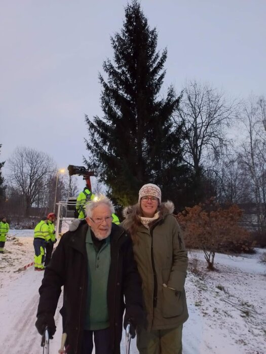 Två personer ler framför ett snötäckt landskap med gran och arbetare i bakgrunden.