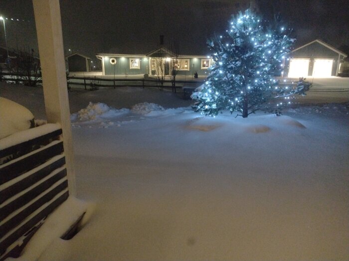 En snöig scen med upplyst julgran vid skymning, bostadshus i bakgrunden, fridfull vinterkväll.