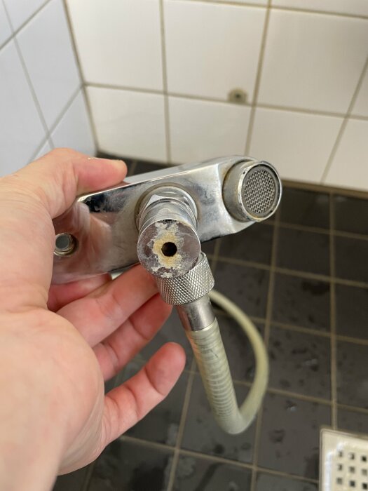 Hand håller en duschslang och duschhuvud med kalkavlagringar på ett badrum med vita kakelväggar.