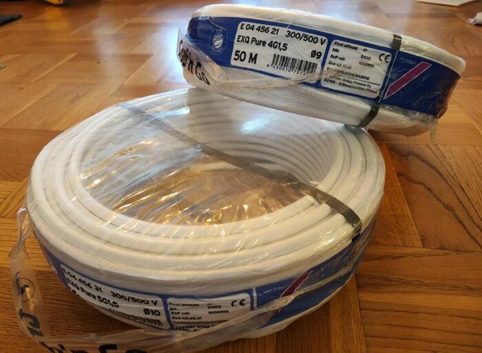 Två rullar vit elektrisk kabel. Förpackningar med teknisk specifikation. Ligga på trägolv.