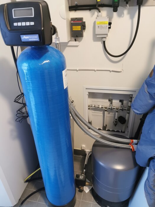 Ett vattenfiltreringssystem med blå behållare, kontrollpanel och röranslutningar i ett tekniskt rum.