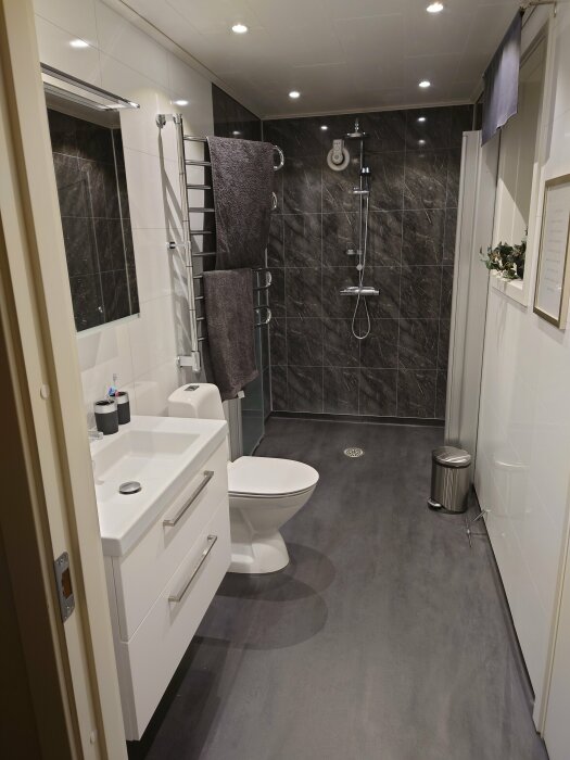 Modernt badrum med dusch, handdukstork, vita skåp, toalett, och gråa toner.