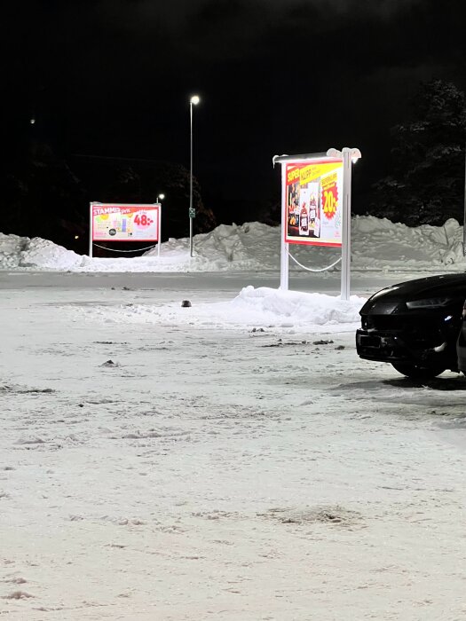 Parkeringsplats nattetid, snötäckt, belyst reklamskyltar, bil i förgrunden, mörk himmel.