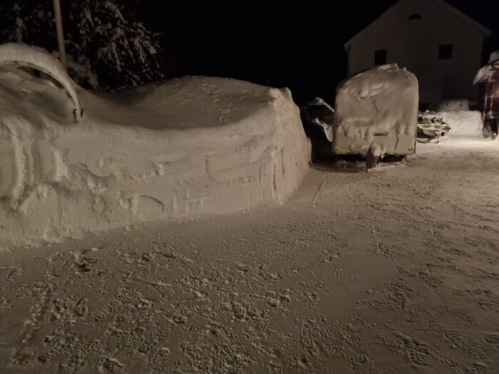 Bilar täckta av snö på parkering nattetid, snödrivor, kall vinter, mörk bakgrund, hus i bakgrunden.
