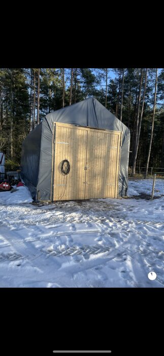 Trätält med presenning i snöigt landskap, skogsbakgrund, verktyg och utrustning synlig på marken.