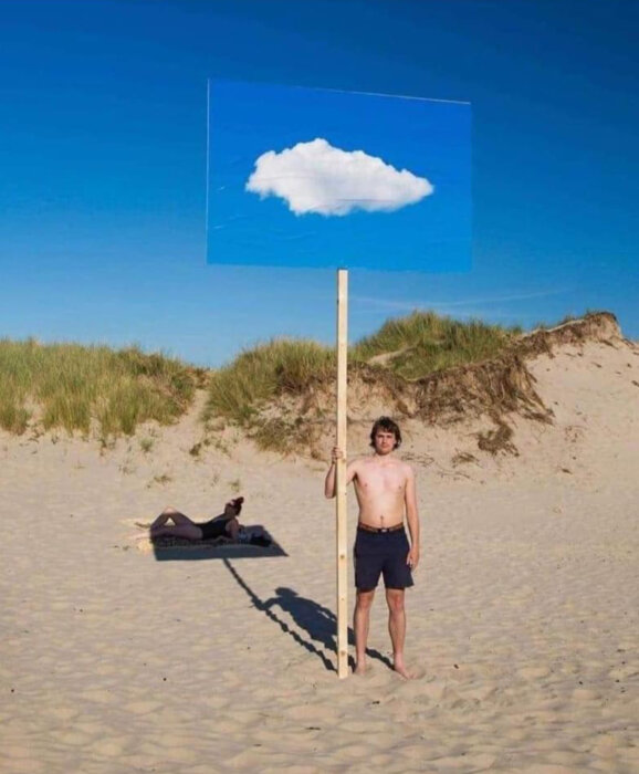 Person håller skylt med moln på stranden, konstnärlig illusion, klar himmel, sanddyner, avslappnad atmosfär.