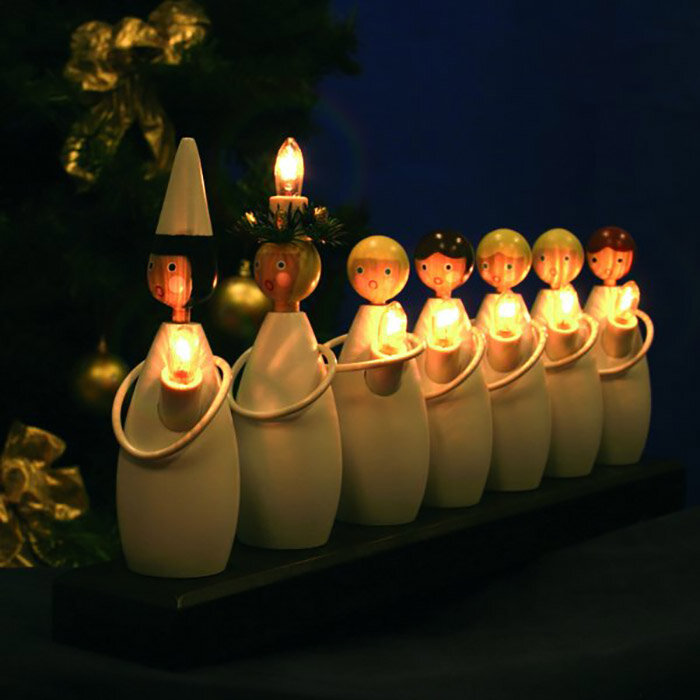 Ljusstake med figurer som håller ljus, juldekoration, mörk bakgrund, julgran syns delvis.