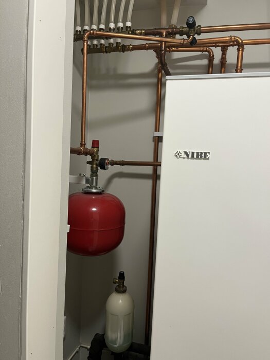 Värmesystem med kopparledningar, expansionskärl och värmepump märkt "NIBE" i ett pannrum eller teknikskåp.