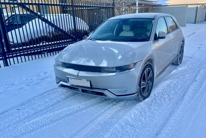 Grå bil parkerad på snötäckt gata, vintertid, dagsljus, spår i snön, staket i bakgrunden.