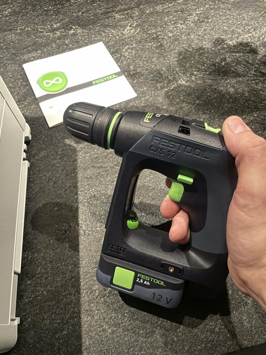 Hand håller en Festool borr/skruvdragare, bord med broschyrer. Svart, grön, 12V, 2.5Ah batteri.