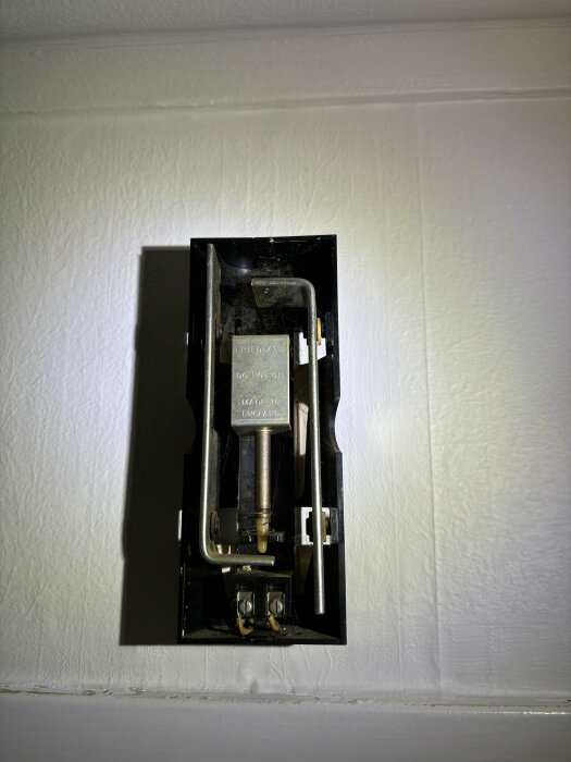 Äldre termisk termostat monterad på vägg, svart och metall, dammig, använd, tillverkad i England.