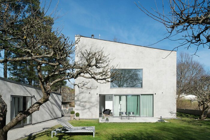 Modernt hus, stora fönster, minimalistisk design, grönt gräs, nakna träd, soligt, klar himmel.