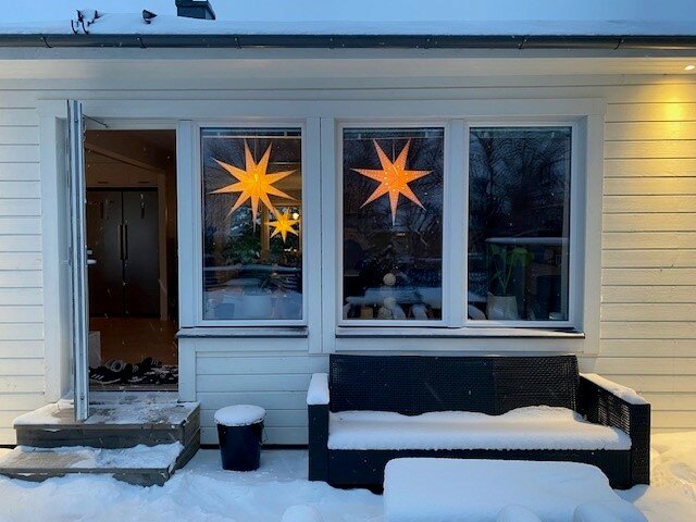 Husfasad med Adventsstjärnor i fönster, öppen dörr, snötäckt bänk och mörklagt landskap.