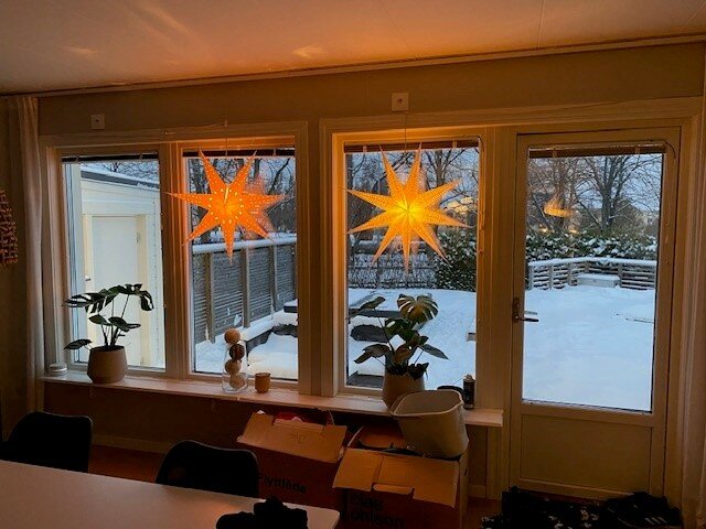 Inomhus vy med fönsterstjärnor, krukväxter och utsikt över snötäckt trädgård vid skymningen.