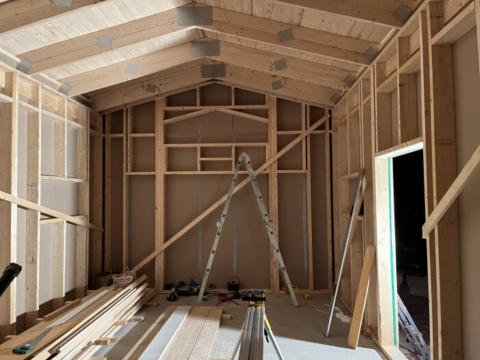 Inomhusbyggarbetsplats med trästomme, brädlager och byggmaterial, högt i tak, halvfärdig konstruktion.