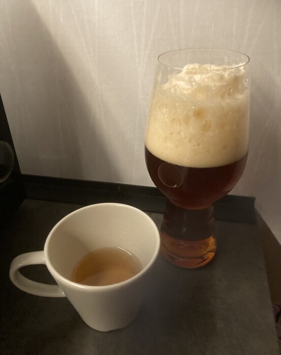 Ölglas med överskummande skum vid sidan av en vit kaffekopp med lite dryck. Skuggor, bänk.