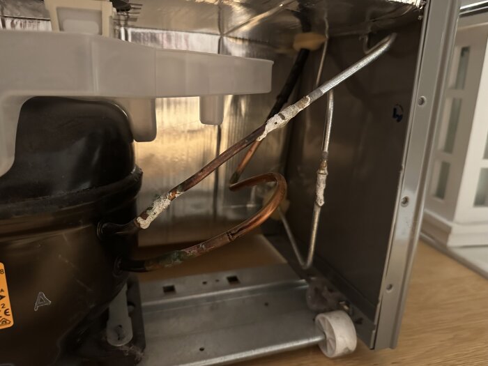 Inre del av en kylskåps baksida, med kompressor och kondensationsrör. Slitage och behov av rengöring synlig.