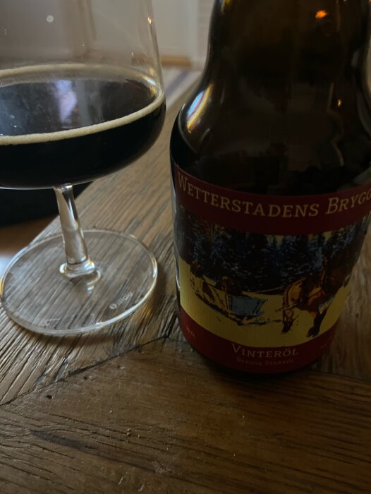 Mörkt öl i glas bredvid ölflaska på träbord. "Vinteröl" etikett, artighet av Wetterstadens Bryggeri.