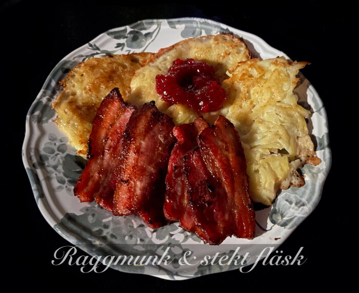 En tallrik med raggmunk, stekt fläsk och lingonsylt på mönstrat porslin mot mörk bakgrund.