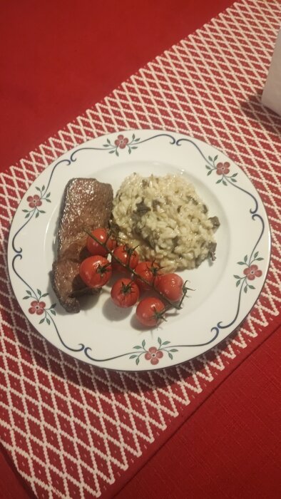 En tallrik med biff, risotto och körsbärstomater på ett röd-vitmönstrat bord.