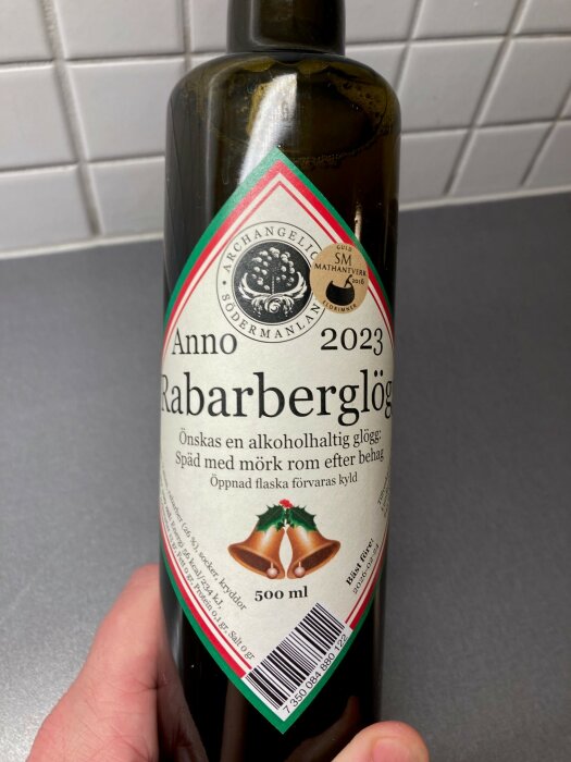 En flaska rabarberglogg, årgång 2023, hålls framför en kaklad vägg. Flaskan är 500 ml.