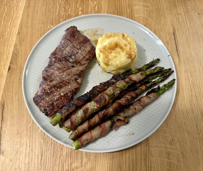 Grillad köttbit, baconlindad sparris, potatisgratäng på en tallrik mot träbakgrund.